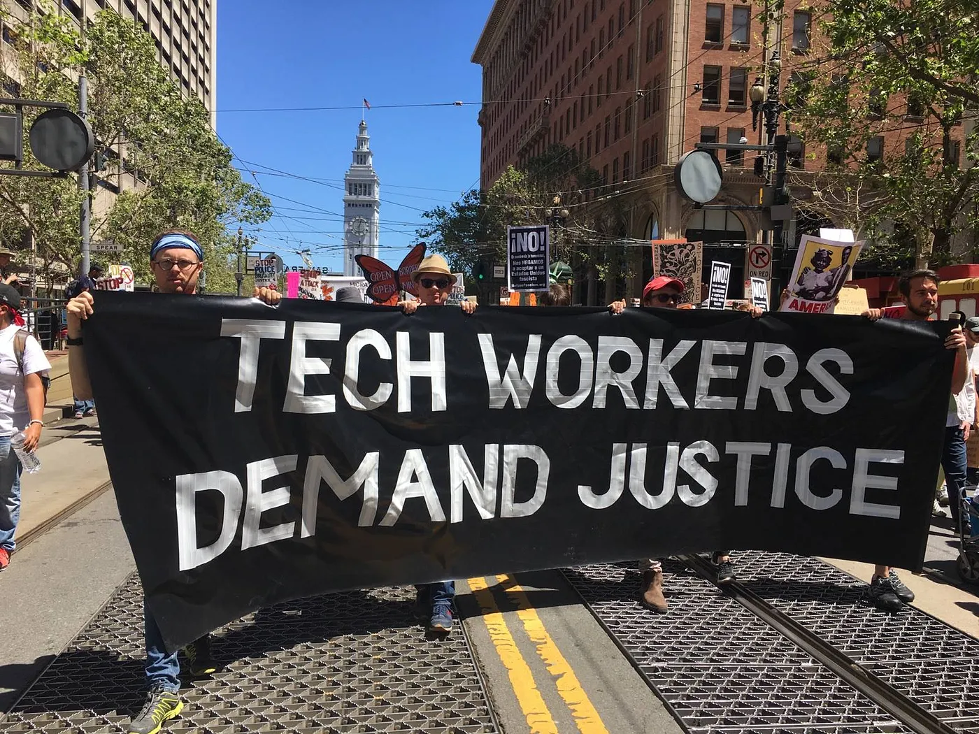 Group of people outside marching while holding a large banner with text that reads "TECH WORKERS DEMAND JUSTICE"